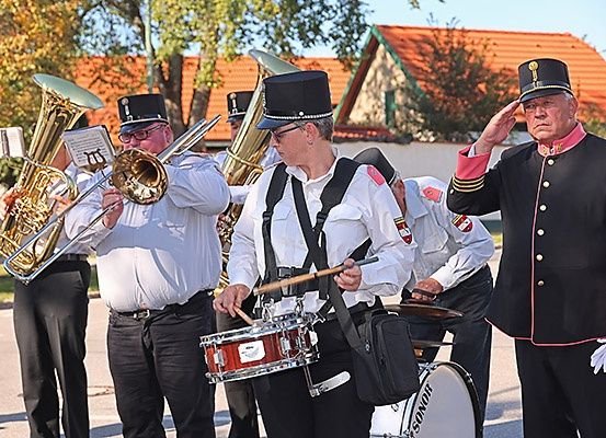 Eine Blaskapelle spielt im Freien, ein Mann salutiert vorne. Sie spielen Instrumente wie Trompete, Posaune und Trommeln.