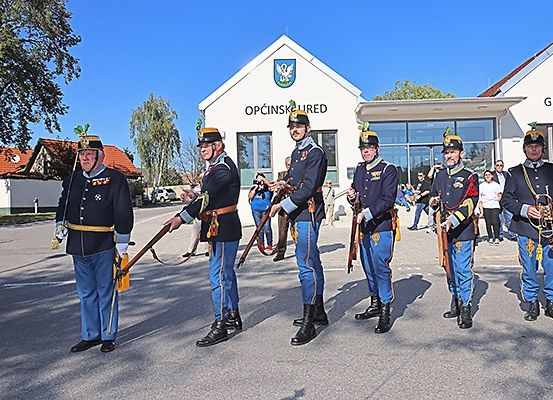 Fünf Männer in Uniform stehen in einer Reihe und halten Gewehre und Trompeten vor einem Gebäude.