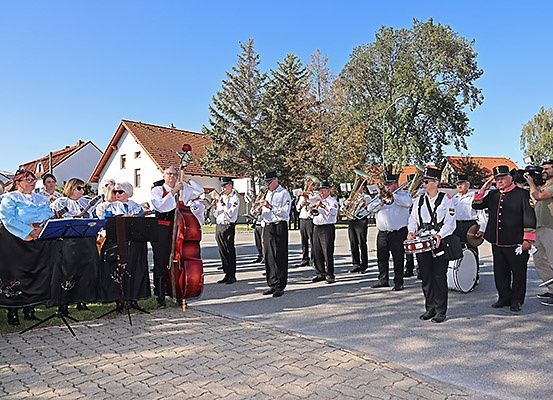 Ein Marschkapelle, bestehend aus Trompeten, Posaunen und Trommeln, spielt auf einer gepflasterten Straße vor Häusern und Bäumen.