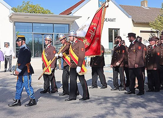 Eine Gruppe uniformierter Personen, möglicherweise Militär, steht vor einem Gebäude. Sie halten Fahnen und tragen Mützen und Handschuhe. Hinter ihnen sind weitere Personen anwesend.