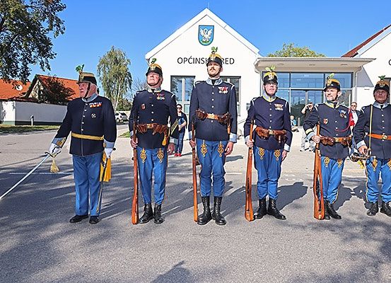 Fünf Männer in Uniform stehen in einer Reihe vor einem Gebäude, jeder hält eine Gewehr und trägt einen Hut. Das Gebäude hat ein Schild, auf dem 'OPCINSKI RED' steht.