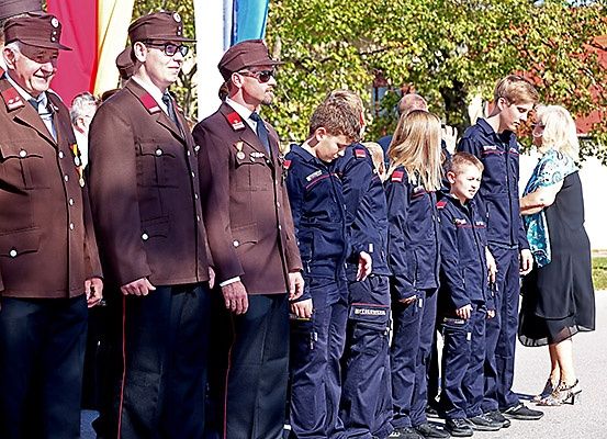 Mehrere Männer in Militäruniformen stehen in einer Reihe, einer trägt eine Brille, ein anderer eine Medaille. Neben ihnen schauen zwei Jungen und eine Frau zu. Dahinter ist eine Flagge und Bäume zu sehen.