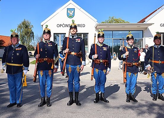 Fünf Männer in Militäruniformen stehen in einer Reihe und halten Gewehre. Sie stehen vor einem Gebäude mit der Aufschrift 'OPCINS UREED'. Dahinter sind verschwommene Figuren von Menschen, möglicherweise bei einer Parade.
