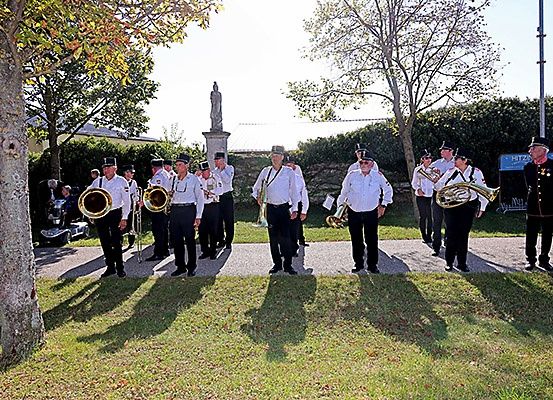 Eine Gruppe von Männern in weißen Uniformen und Hüten spielt im Freien und trägt Blechblasinstrumente. Der Ort ist auf einer Rasenfläche mit Bäumen und einer Statue im Hintergrund.