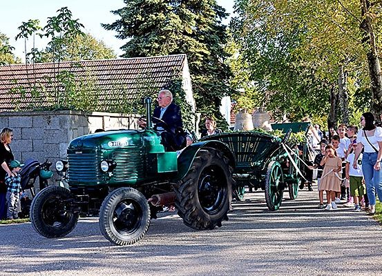 Ein älterer Mann fährt einen grünen Traktor mit Anhänger, gefolgt von mehreren Kindern und Erwachsenen. Die Szene spielt sich im Freien ab, mit Bäumen und einem Gebäude im Hintergrund.