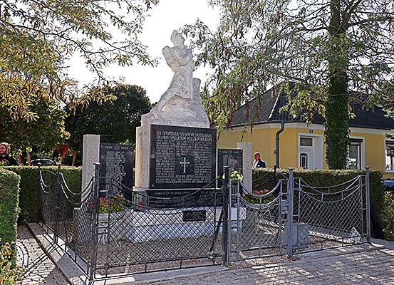 Eine Statue einer Person ist von einem Metallzaun umschlossen. Hinter dem Zaun befindet sich ein Denkmal mit einem Kreuz und Namen auf einer schwarzen Tafel. Eine Person steht in der Nähe des Denkmals. Es gibt ein Haus mit gelben Wänden und ein Auto, das davor geparkt ist.
