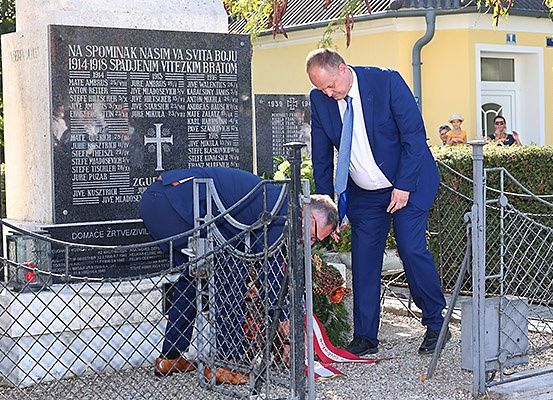 Zwei Männer in Anzügen, einer mit einem Kranz, stehen an einem Denkmal, einer verneigt sich. Dahinter steht eine Tafel mit Namen, und im Hintergrund befindet sich ein Gebäude.