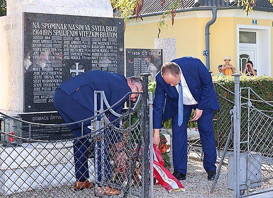 Zwei Männer in Anzügen legen einen Kranz an einem Denkmal nieder. Dahinter steht ein Gedenkstein mit Namen und Daten, ein Zaun und ein Haus im Hintergrund.