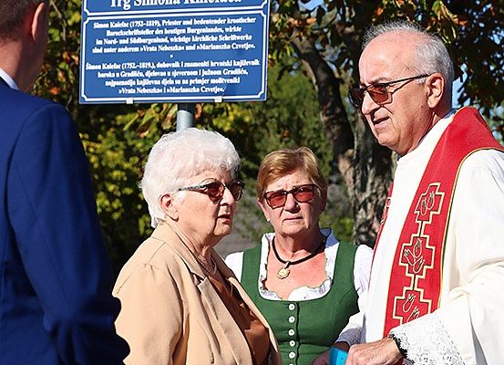 Eine Gruppe von Menschen steht vor einem blauen Schild, einer trägt eine weiß-rote Robe.