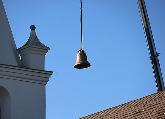 Eine Glocke hängt von einem Kran in der Nähe des Daches eines Kirchengebäudes. Das Gebäude hat ein spitzes Dach und ein Fenster an der Seite.