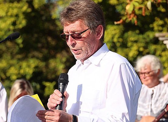 Ein Mann mit Sonnenbrille und weißem Hemd spricht in ein Mikrofon. Dahinter sind verschwommene Personen zu sehen, darunter eine Frau mit einer Gitarre.