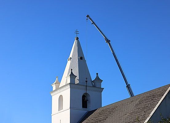 Ein Kran hebt eine Glocke zur Spitze eines Kirchturms unter einem klaren blauen Himmel.