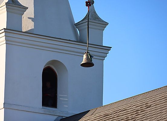 Eine Glocke hängt an einem Turm einer Kirche, wahrscheinlich zum Signalisieren oder zur Zeitanzeige. Das Gebäude hat eine weiße Außenfassade und ein braunes Dach.