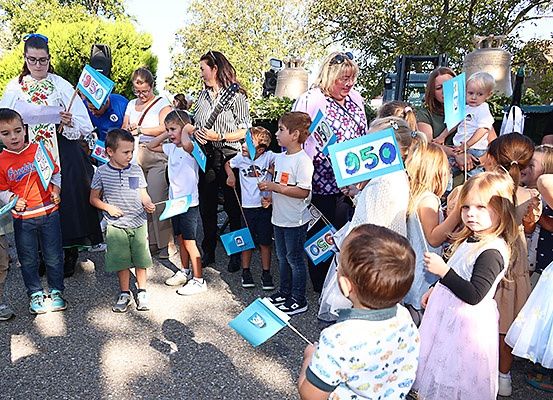Eine Gruppe von Kindern und Erwachsenen versammelt sich im Freien und hält blaue Flaggen mit Nummern. Sie scheinen an einer Veranstaltung teilzunehmen. Einige tragen Freizeitkleidung, andere Kleider. Hinter ihnen sind Bäume und ein Glockenturm zu sehen.