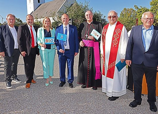 Eine Gruppe von Menschen, darunter Geistliche und Laien, steht lächelnd für ein Foto vor einer Kirche. Einige halten blaue Fahnen mit der Nummer 950.