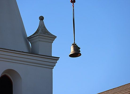 Eine Glocke hängt an einem Metallseil an einem Kirchturm gegen einen klaren blauen Himmel.