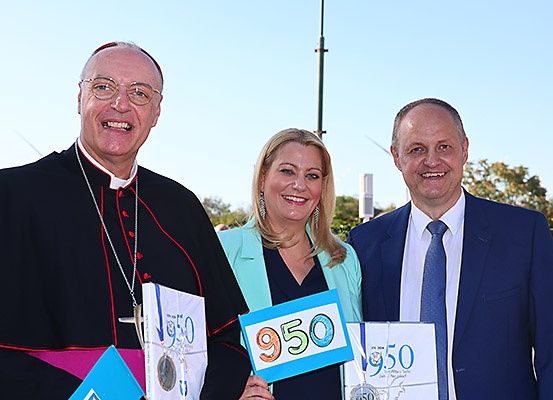 Drei Menschen stehen lächelnd nebeneinander und halten Schilder mit der Nummer 950 in der Hand, vor einer malerischen Kulisse.