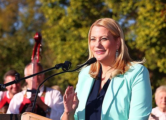 Eine Frau spricht in ein Mikrofon bei einer Veranstaltung im Freien. Hinter ihr spielen Musiker Instrumente, und im Hintergrund sitzen Leute. Bäume und ein klarer Himmel sind sichtbar.