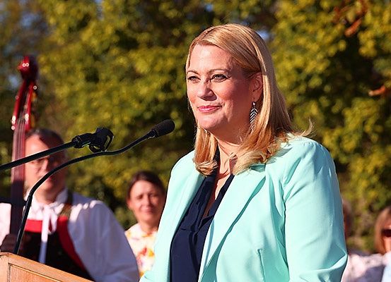 Eine Frau steht an einem Rednerpult mit Mikrofon, das einen hellblauen Blazer trägt. Hinter ihr spielt ein Mann in traditioneller Kleidung Cello, und zwei Frauen schauen zu. Bäume und Grünflächen sind im Hintergrund.