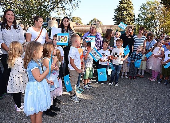 Eine Gruppe von Menschen, darunter Kinder und Erwachsene, steht draußen. Sie halten blaue Schilder mit Nummern, möglicherweise an einer Veranstaltung teilnehmend. Die Kinder tragen Kleider und Turnschuhe.