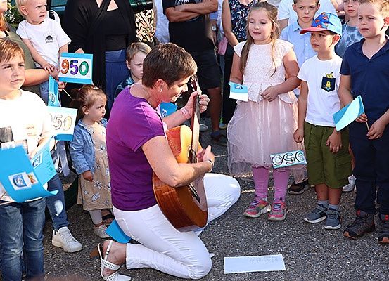 Eine Frau kniet mit einer Gitarre und singt für eine Gruppe von Kindern und Erwachsenen, einige halten Schilder mit Zahlen.