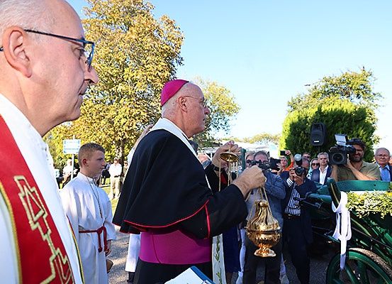 Eine religiöse Prozession mit einem Priester in einem rosa-schwarzen Gewand, der einen goldenen Kelch hält, umgeben von Menschen und Grünpflanzen.