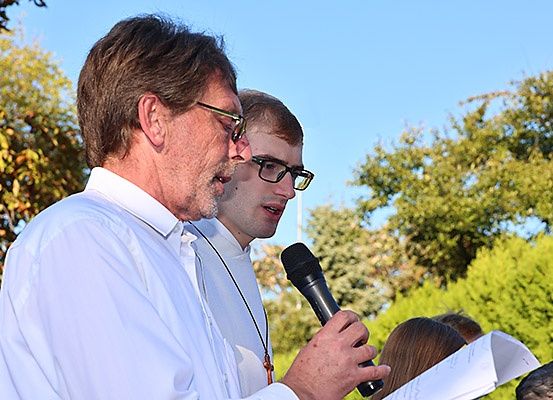 Ein Mann in einem weißen Hemd hält ein Mikrofon und spricht, während ein anderer Mann mit einer Brille und einer Halskette hinter ihm steht. Sie sind draußen mit Bäumen und einem klaren Himmel.