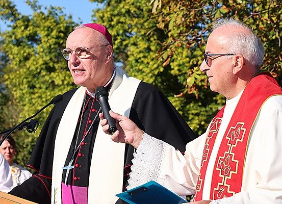 Zwei Männer in Klerikerroben sprechen an einem Podium mit Mikrofon, einer hält ein Buch. Bäume und eine Frau im Hintergrund.