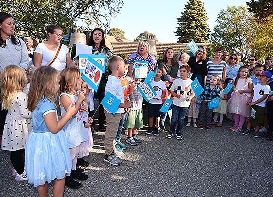 Eine Gruppe von Kindern und Erwachsenen steht im Freien und hält blaue Flaggen mit Zahlen. Einige tragen Brillen, Sneaker und Kleider. Eine Frau in der Mitte schreit. Bäume und ein Gebäude sind im Hintergrund.