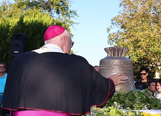 Ein Priester mit roter Zucchetto hält eine Glocke bei einer Veranstaltung im Freien. Dahinter beobachtet eine Menge.
