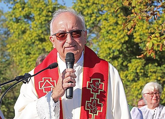 Ein Priester in weißer und roter Robe mit einem Kreuz darauf spricht in ein Mikrofon. Dahinter sind verschwommene Menschen. Bäume sind im Hintergrund.