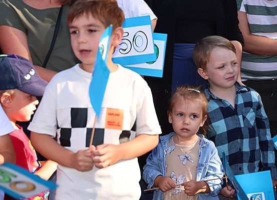 Eine Gruppe von Kindern, die blaue Flaggen mit Nummern halten, steht in einer Reihe, einige tragen Mützen und T-Shirts, während andere zuschauen, einschließlich eines Mädchens in einer Jeansjacke.