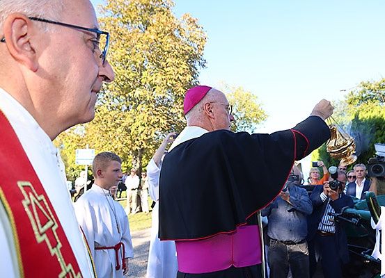 Eine religiöse Zeremonie im Freien mit einem Bischof, Messdienern und Zuschauern. Der Bischof erhebt seine Hände in einer Geste. Einige Teilnehmer machen Fotos.
