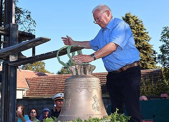 Ein älterer Mann steht auf einer Glocke und justiert sie mit einem Seil, während Leute in der Nähe zusehen. Hinter ihm ist ein Haus mit einem roten Dach sichtbar.