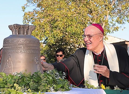 Ein Bischof in traditioneller Kleidung hält einen Kelch bei einer Zeremonie, steht neben einer Glocke mit religiösen Inschriften und ist von Menschen und Grünpflanzen umgeben.