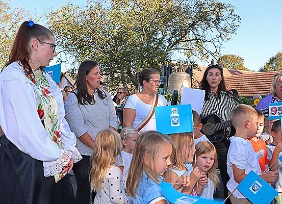 Eine Gruppe von Erwachsenen und Kindern versammelt sich in einer Außenumgebung, möglicherweise für eine öffentliche Veranstaltung. Einige halten blaue Banner, und eine Frau mit Brille steht prominent.
