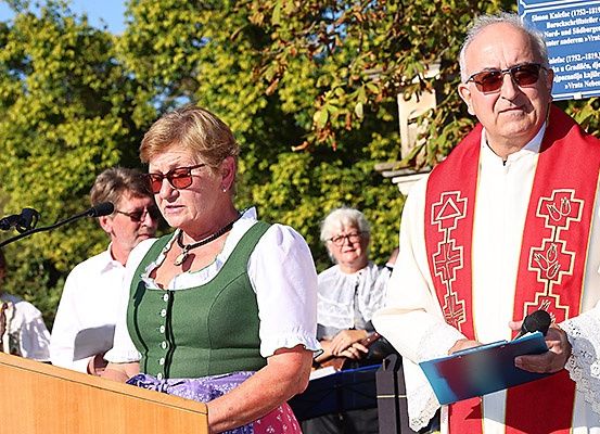 Eine Gruppe von Menschen steht in einer Outdoor-Umgebung, wahrscheinlich für eine Zeremonie. Eine Frau steht an einem Podium, während ein Mann neben ihr eine Mappe und ein Mikrofon hält. Hinter ihnen beobachten mehrere Personen. Der Hintergrund zeigt Bäume und ein blaues Schild.