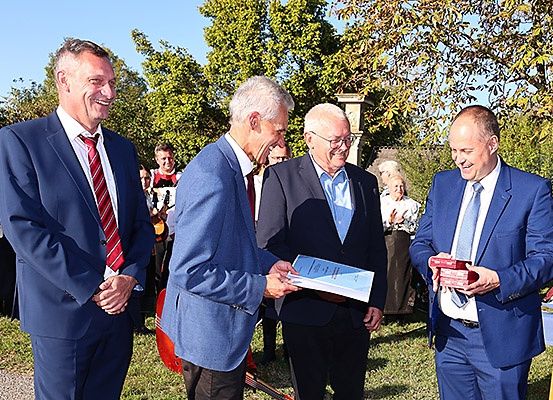 Vier Männer in Anzügen sind draußen. Zwei Männer tauschen Geschenke aus, während die anderen zusehen. Bäume und Menschen sind im Hintergrund.