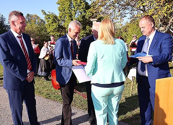 Eine Gruppe formell gekleideter Menschen steht draußen, wahrscheinlich bei einer Veranstaltung. Ein Mann hält ein Band, während ein anderer Mann ein Dokument liest. Bäume und eine Rasenfläche sind im Hintergrund zu sehen.