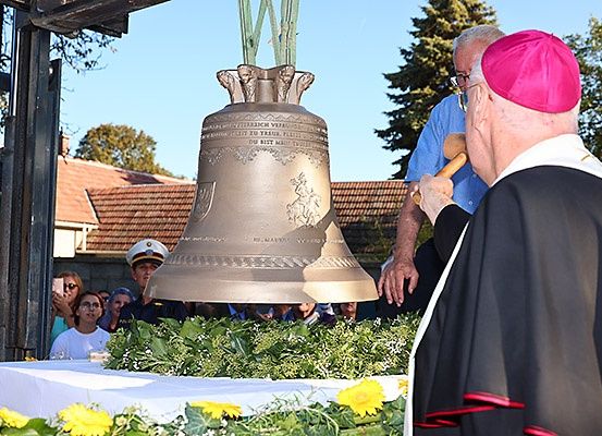 Eine große Glocke wird herabgelassen, während ein Priester einen Hammer hält. Ein Mann in einem blauen Hemd schaut zu, und andere beobachten im Hintergrund.