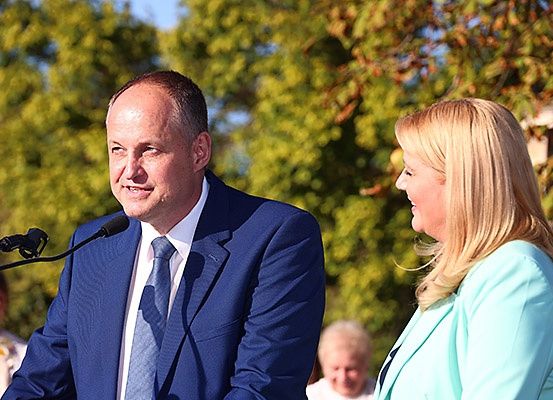 Ein Mann und eine Frau stehen an einem Podium, der Mann spricht in ein Mikrofon und die Frau schaut ihn an. Hinter ihnen sind Bäume und Menschen im Hintergrund.