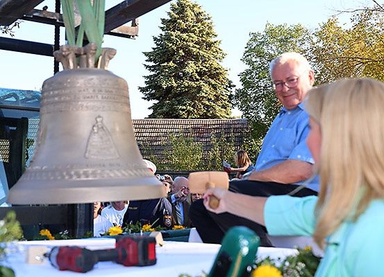 Ein älterer Mann und eine Frau sitzen an einem Tisch mit Werkzeugen. Ein großer Glockenturm ist hinter ihnen, und eine Menge Leute steht im Hintergrund.