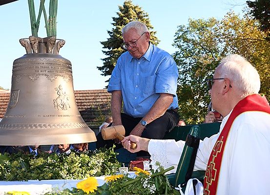 Zwei Männer sind bei einer Kirchenveranstaltung. Einer hält einen Hammer und der andere ist in einer Robe. Eine Glocke steht hinter ihnen.