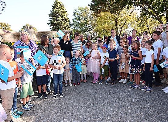 Eine Gruppe von Kindern und Erwachsenen steht zusammen und hält blaue Flaggen mit Zahlen. Sie scheinen ein Ereignis auf einem Kiesweg zu feiern.