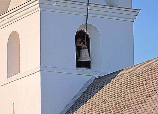 Eine Person befindet sich in der Glockenkammer eines Turms und hält eine mit einer Glocke verbundene Seil. Der Turm hat ein weißes Äußeres und ein braunes Dach.