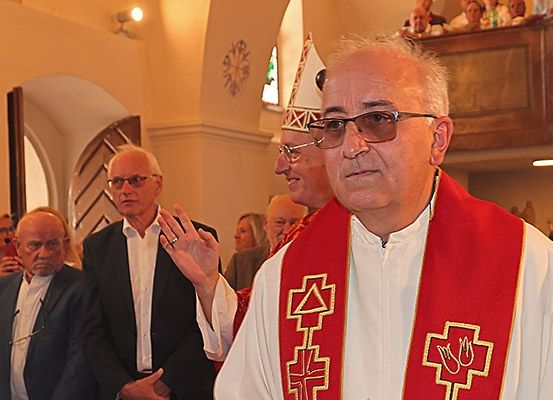 Ein Priester steht in einer Kirche mit einem Kreuz auf seinem Gewand und blickt nach vorne. Hinter ihm stehen mehrere Menschen, und einige sitzen auf einer Galerie.