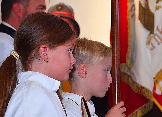 Zwei Kinder in weißen Uniformen stehen nebeneinander und halten einen Fahnenmast. Hinter ihnen steht ein Erwachsener, und eine Flagge hängt hinter den Kindern.