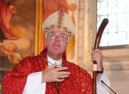 Ein Bischof spricht in ein Mikrofon und hält einen Holzstab, gekleidet in rote und weiße Gewänder.