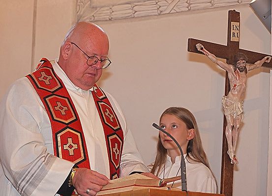 Ein Priester steht hinter einem Pult und liest aus einem Buch, ein Mädchen neben ihm hört aufmerksam zu. Dahinter ist ein Kruzifix mit INRI auf dem Kreuz an der Wand angebracht.