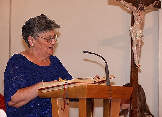 Eine ältere Frau mit Brille liest aus einem Buch an einer Kanzel in einer Kirche, mit einem Kreuz und Jesus am Kreuz hinter ihr. Ein Mikrofon steht vor ihr. Ein Mädchen sitzt im Hintergrund.